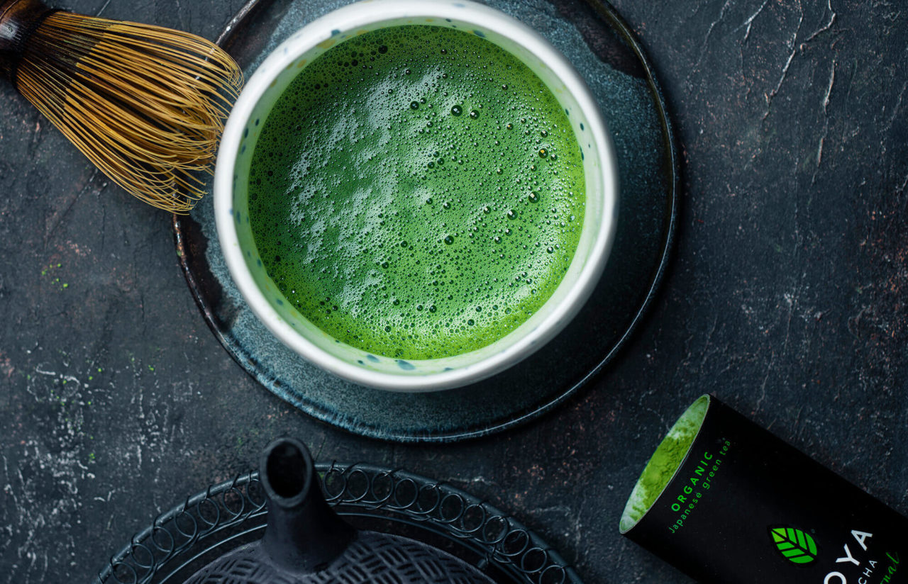 Een kom vers opgeklopte matcha met een bamboe garde, naast een zwart blik Moya matcha